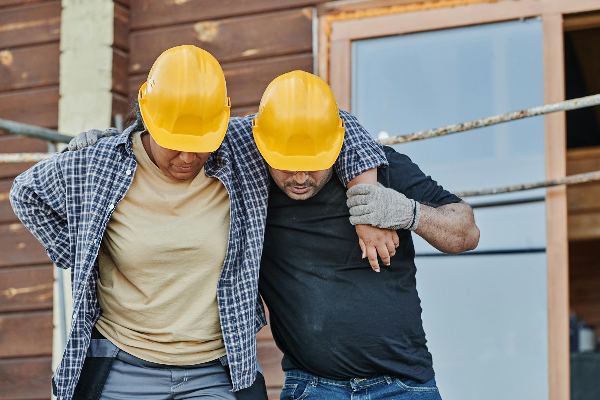 two construction workers in hard hats, one of which appears injured and is being assisted by the other.